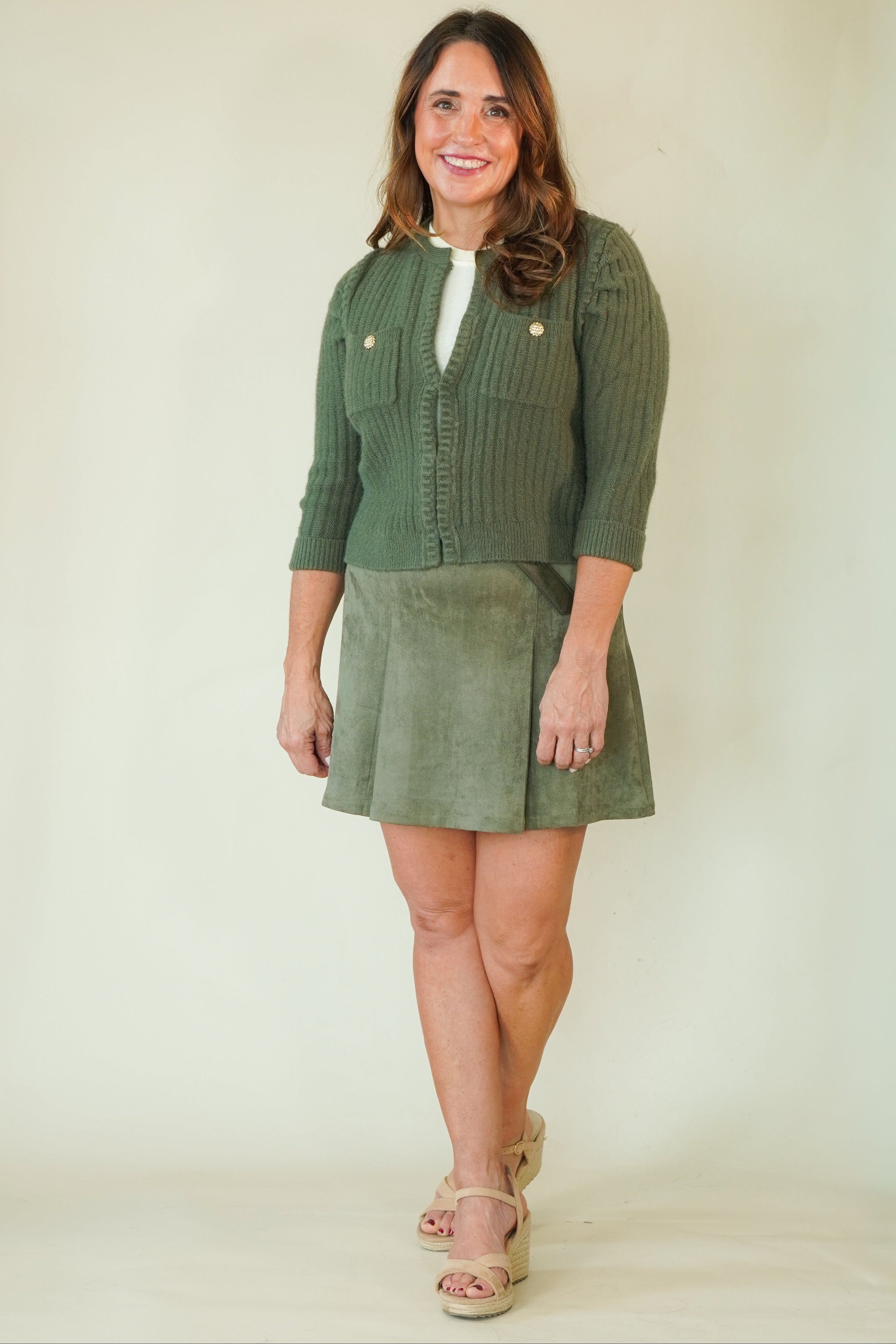 A woman modeling a two-piece olive green outfit consisting of a ribbed knit cardigan with gold buttons and a matching corduroy skirt, paired with beige platform sandals, against a plain cream background.