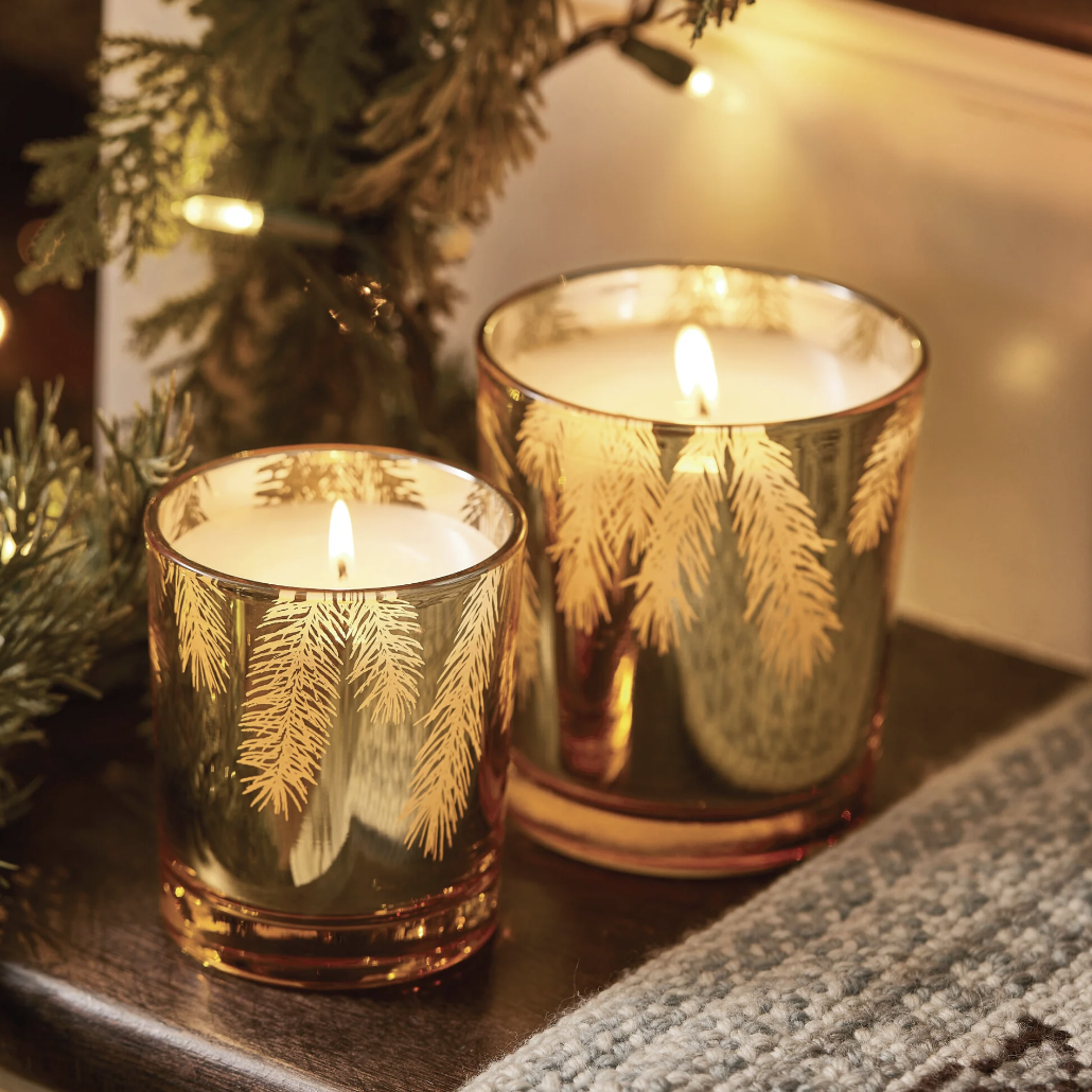 Two lit candles in decorative glass holders with a Christmas tree in the background.