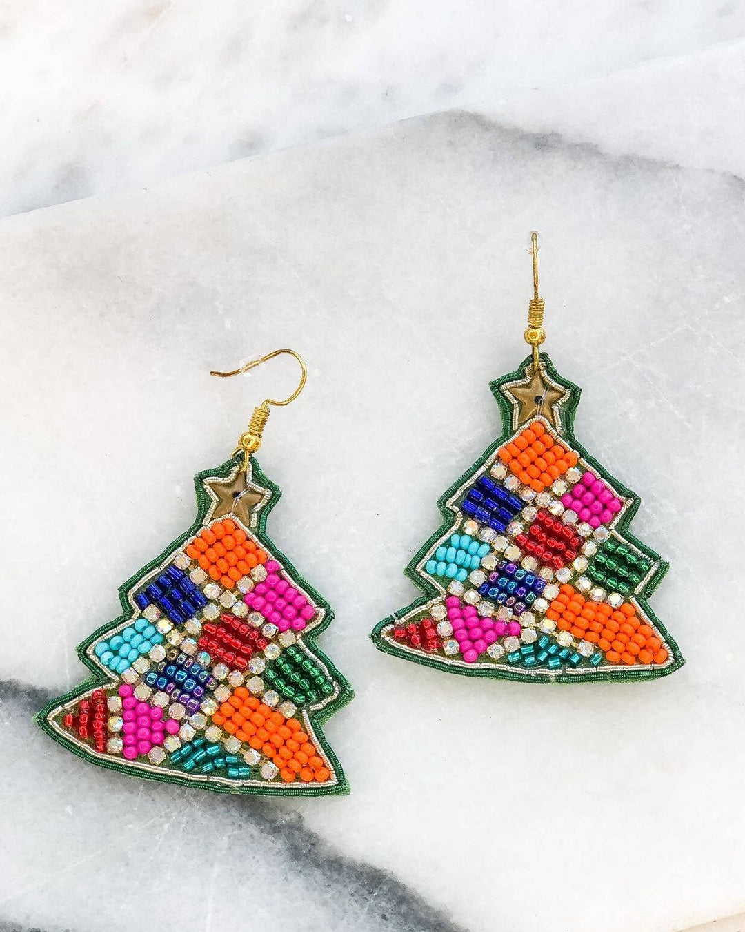 A pair of beaded earrings designed to resemble a Christmas tree, with red, green, blue, and yellow colors on a white background, displayed on a marble surface.