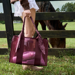 A person standing in a grassy field beside a wooden fence holds a sheer burgundy tote bag with a matching fabric bandana-style handle. A horse is visible behind the fence in the background.