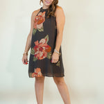 A woman modeling a sleeveless high-neck mini dress with a dark gray base and a large floral print in orange, red, and green, paired with beige wedge sandals against a plain cream background.