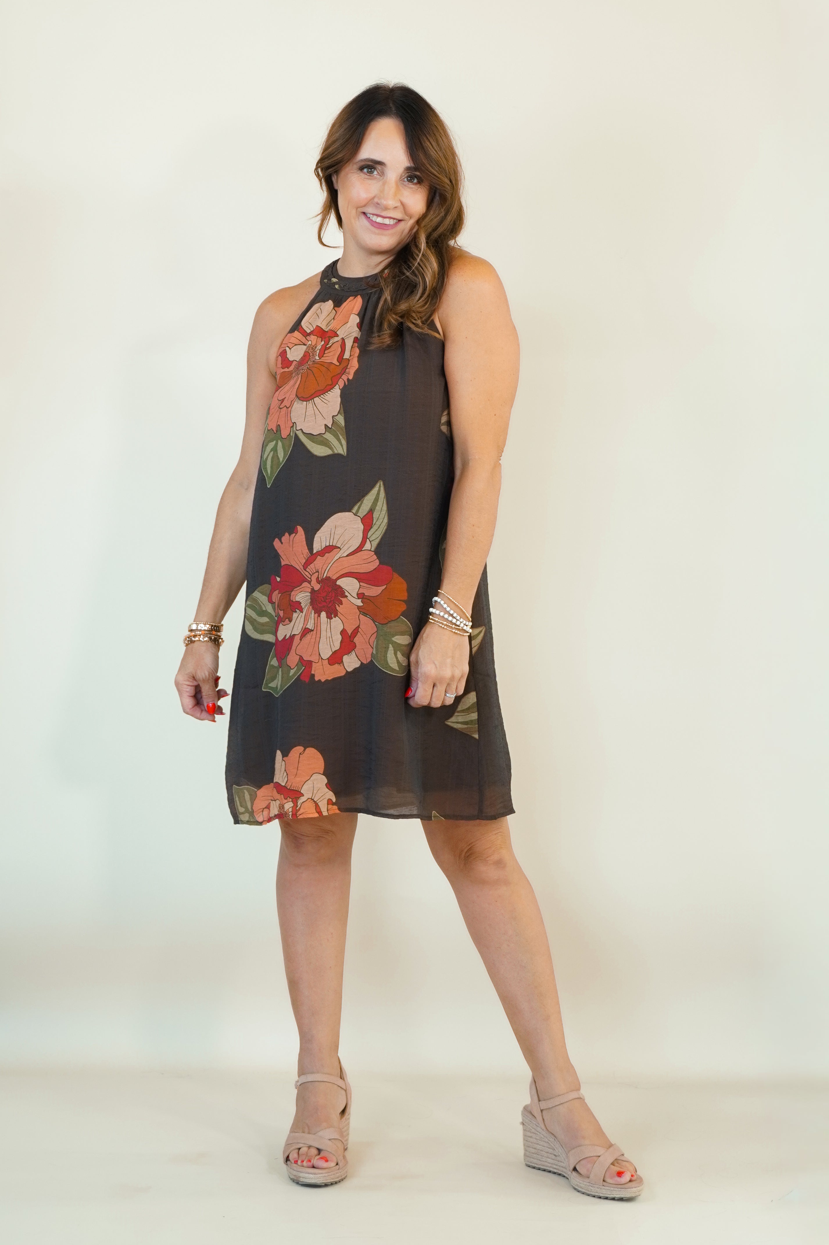 A woman modeling a sleeveless high-neck mini dress with a dark gray base and a large floral print in orange, red, and green, paired with beige wedge sandals against a plain cream background.