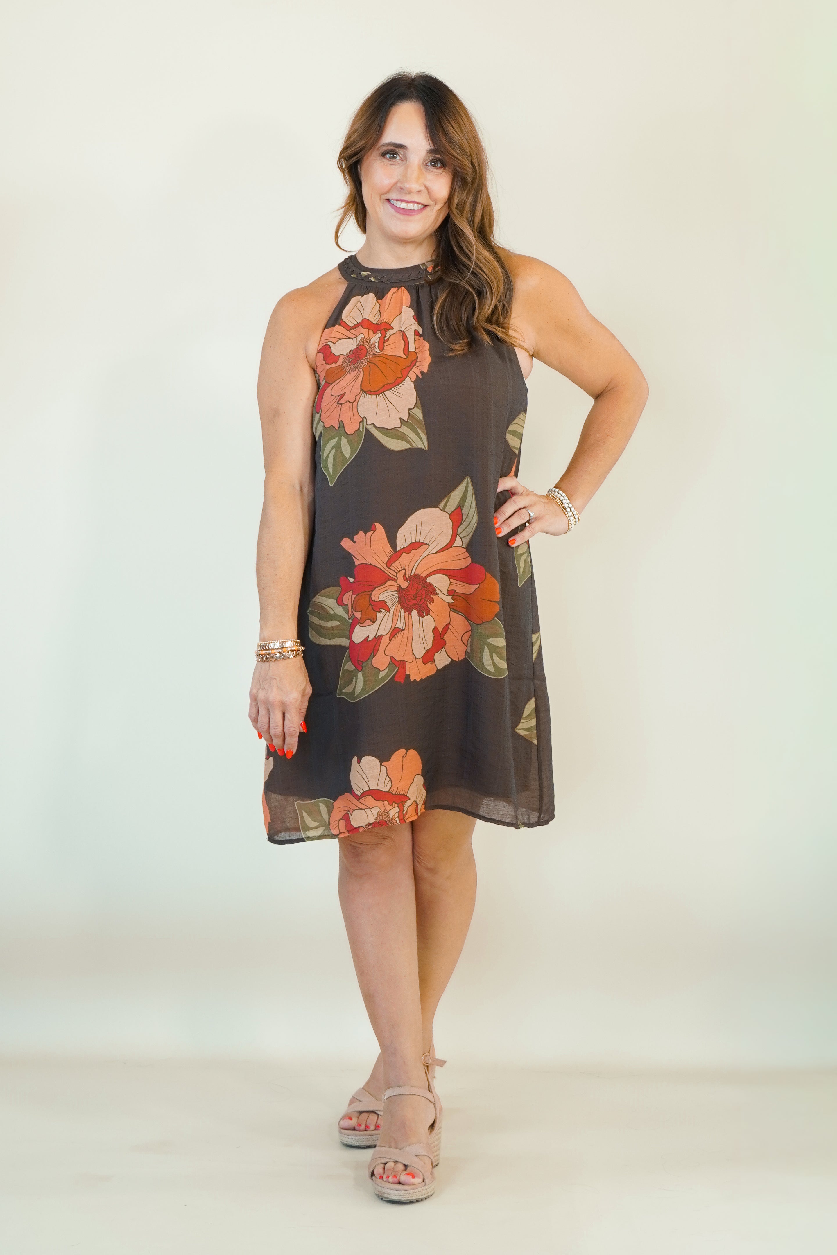 A woman modeling a sleeveless halter-neck shift dress with a dark brown base and a large orange, peach, and green floral print, paired with beige strappy sandals against a plain light beige studio background.