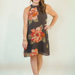 A woman modeling a sleeveless halter-neck shift dress with a dark brown base and a large orange, peach, and green floral print, paired with beige strappy sandals against a plain light beige studio background.