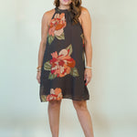 A woman modeling a sleeveless halter-neck mini dress with a dark brown base and a large floral print in orange, red, and green, paired with beige wedge sandals against a plain off-white studio background.