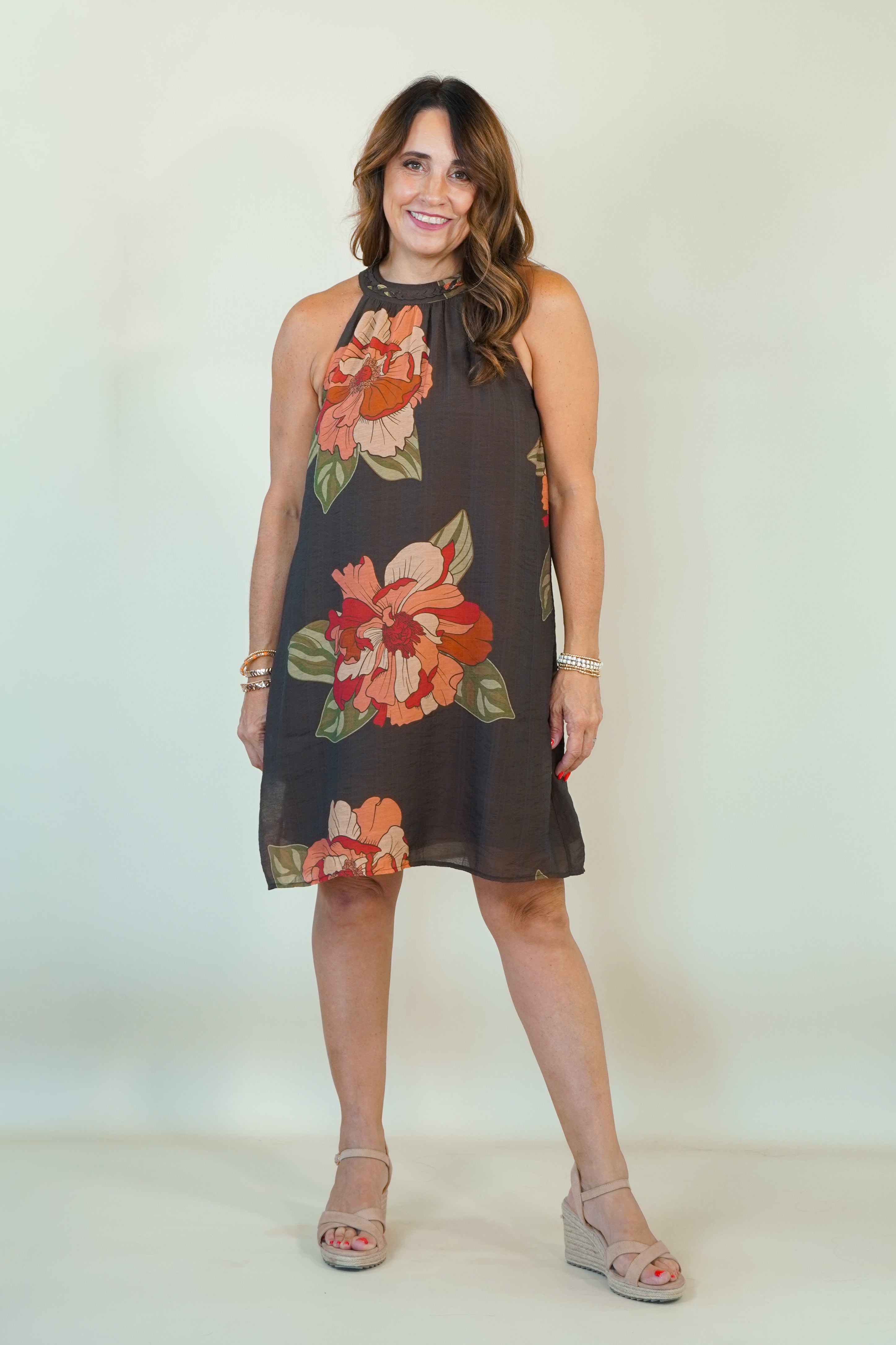 A woman modeling a sleeveless halter-neck mini dress with a dark brown base and a large floral print in orange, red, and green, paired with beige wedge sandals against a plain off-white studio background.