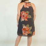 A woman viewed from behind wearing a sleeveless halter-neck dress with a dark charcoal base and a large floral print in orange, pink, and green, paired with beige wedge sandals against a plain off-white studio background.
