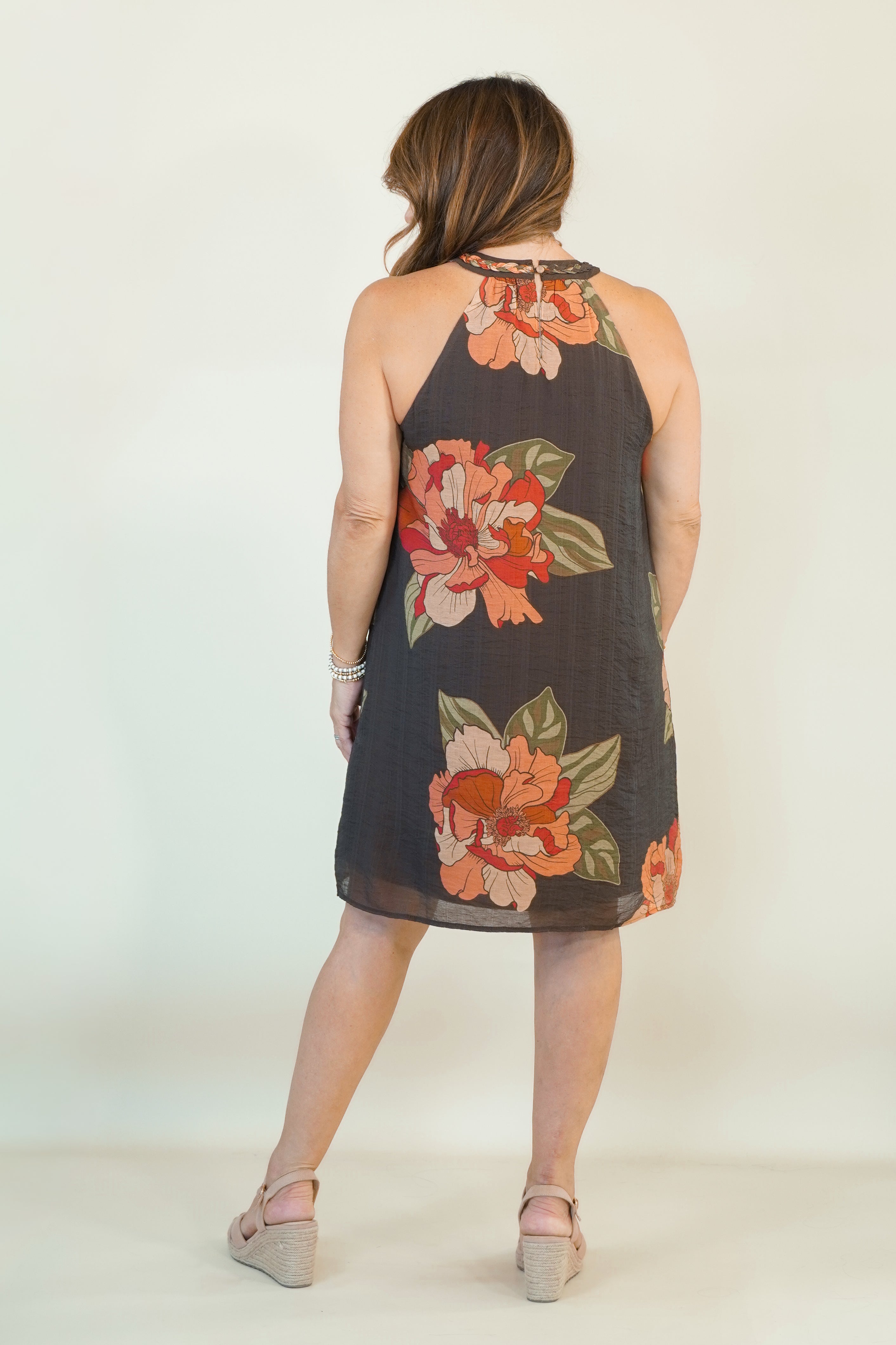 A woman viewed from behind wearing a sleeveless halter-neck dress with a dark charcoal base and a large floral print in orange, pink, and green, paired with beige wedge sandals against a plain off-white studio background.