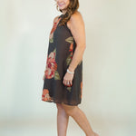 A woman modeling a sleeveless dark brown shift dress with a red and green floral print, paired with beige wedge sandals, against a plain off-white studio background.