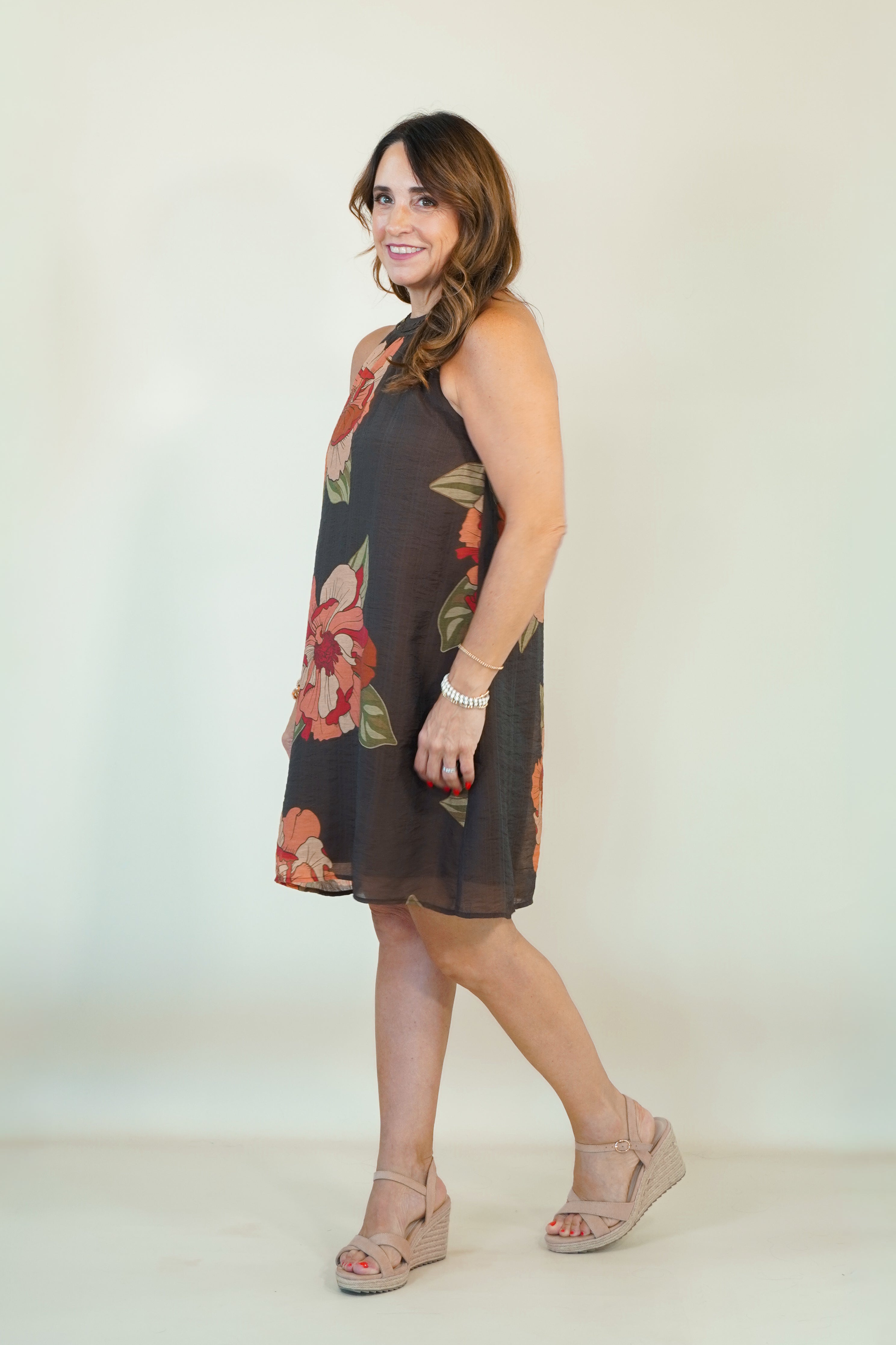 A woman modeling a sleeveless dark brown shift dress with a red and green floral print, paired with beige wedge sandals, against a plain off-white studio background.