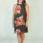 A woman modeling a sleeveless high-neck A-line dress with a dark brown base and a large floral print in red, orange, and green, standing against a plain off-white studio background.