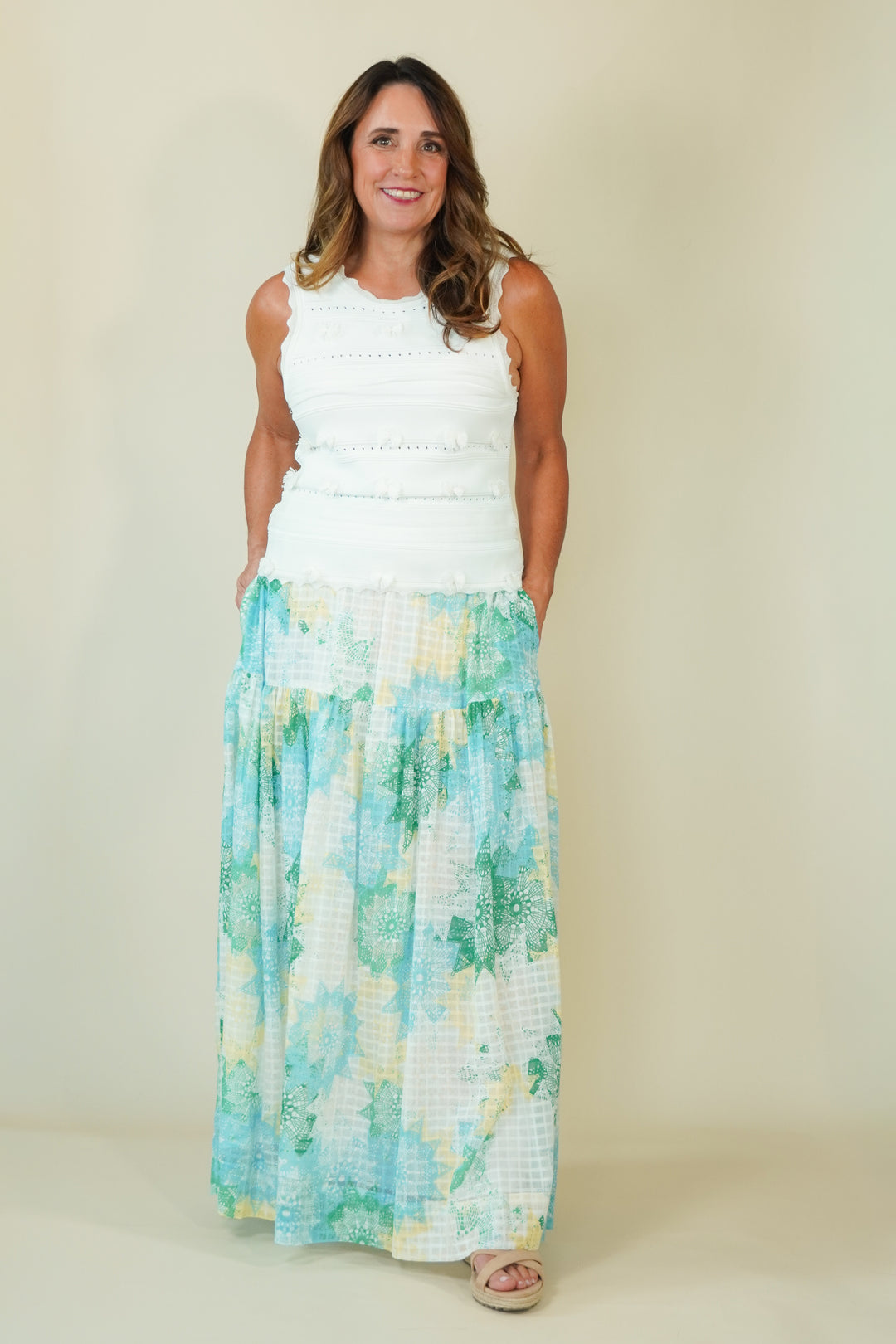 A woman modeling a sleeveless white crop top and a long tiered maxi skirt with a blue, green, and white watercolor floral pattern, standing against a plain beige background.