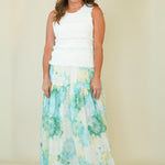 A woman modeling a sleeveless white crop top and a long tiered maxi skirt with a green and blue floral print on a white background, standing against a solid beige backdrop.