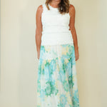 A woman modeling a sleeveless maxi dress with a white textured bodice and a flowing skirt featuring a light blue, green, and yellow floral pattern, set against a plain beige studio background.
