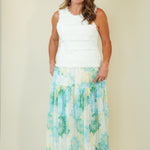 A woman modeling a sleeveless maxi dress with a white textured top and a flowing skirt featuring a green, blue, and yellow tropical floral pattern, set against a plain beige studio background.