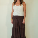 A woman modeling a sleeveless white knit top with a floral embroidery pattern and a long, tiered brown skirt, standing against a plain light beige wall.