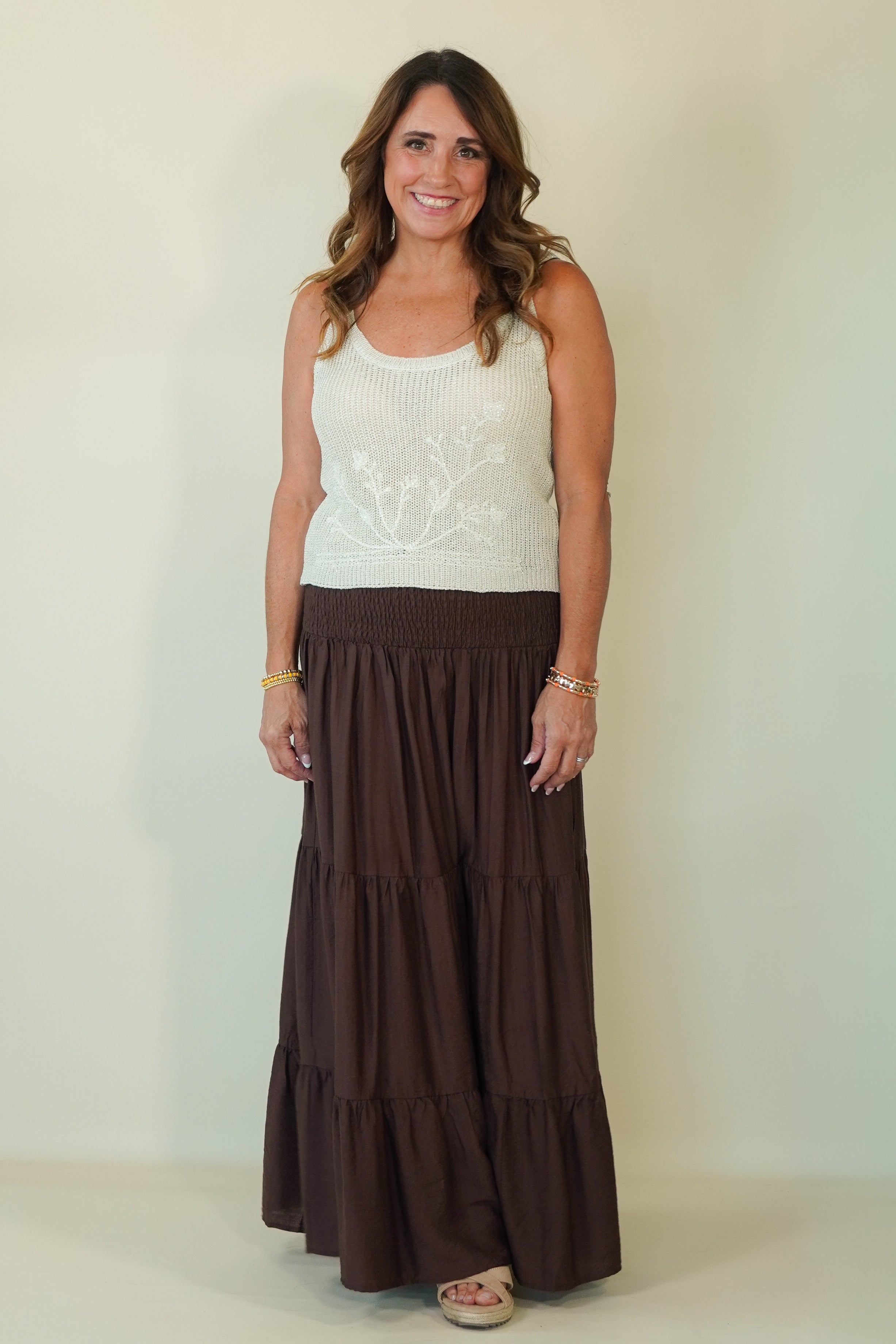 A woman modeling a sleeveless white knit top with a floral embroidery pattern and a long, tiered brown skirt, standing against a plain light beige wall.