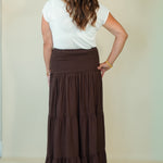 A woman shown from the back wearing a white short-sleeve top and a long brown tiered maxi skirt, standing against a plain light beige wall.