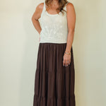 A woman modeling a sleeveless white knit top with a white floral embroidery on the chest, paired with a long brown tiered maxi skirt, standing against a plain beige background.