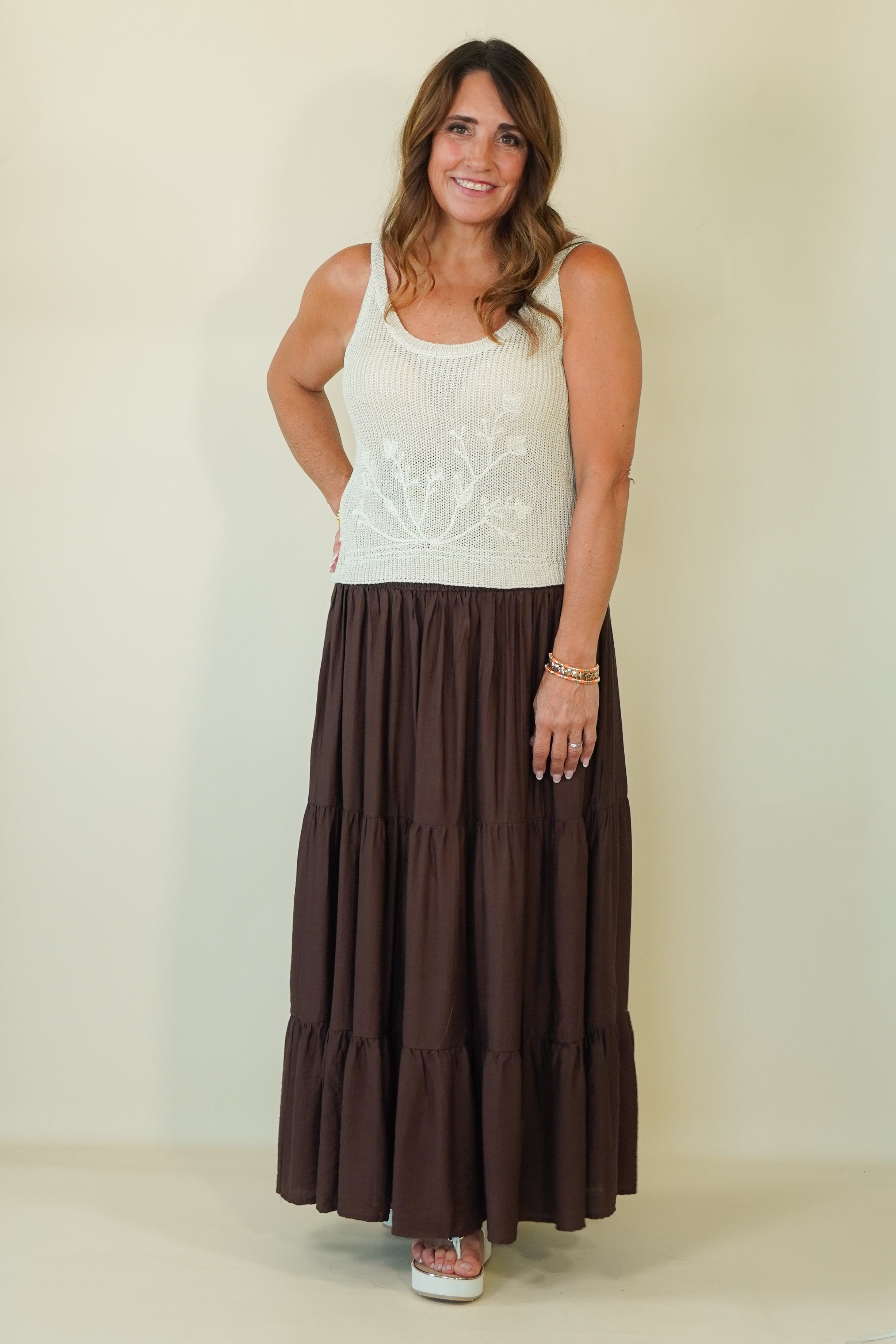 A woman modeling a sleeveless white knit top with a white floral embroidery on the chest, paired with a long brown tiered maxi skirt, standing against a plain beige background.
