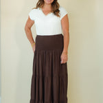 A woman modeling a white short-sleeve V-neck top paired with a long, dark brown tiered maxi skirt, standing against a plain beige studio background.