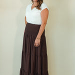 A woman modeling a white short-sleeve V-neck top paired with a long, dark brown tiered maxi skirt, standing against a plain beige studio background.