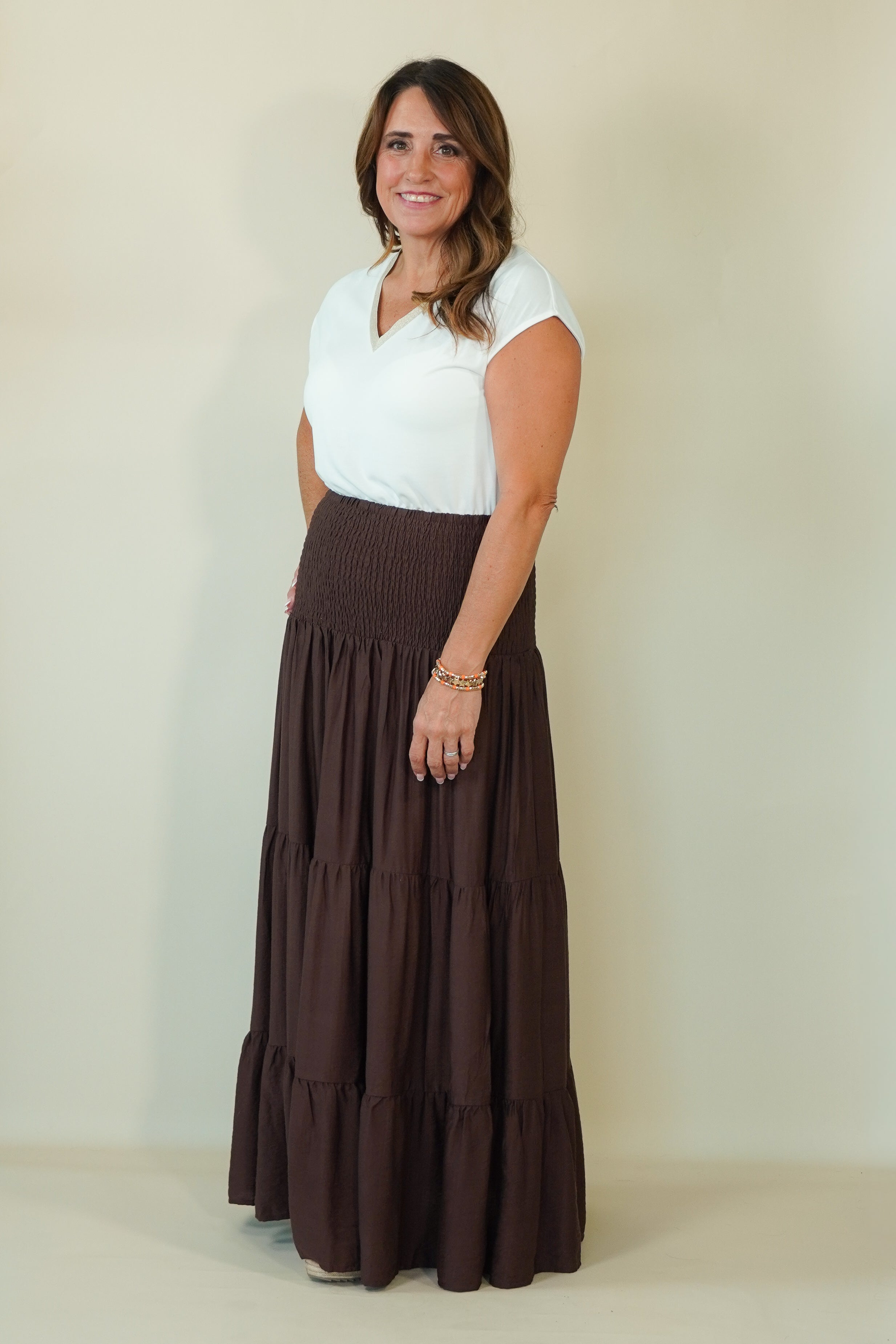 A woman modeling a white short-sleeve V-neck top paired with a long, dark brown tiered maxi skirt, standing against a plain beige studio background.