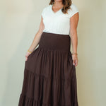 A woman modeling a white short-sleeve V-neck top and a high-waisted, dark brown tiered maxi skirt with side pockets, standing against a plain beige studio background.