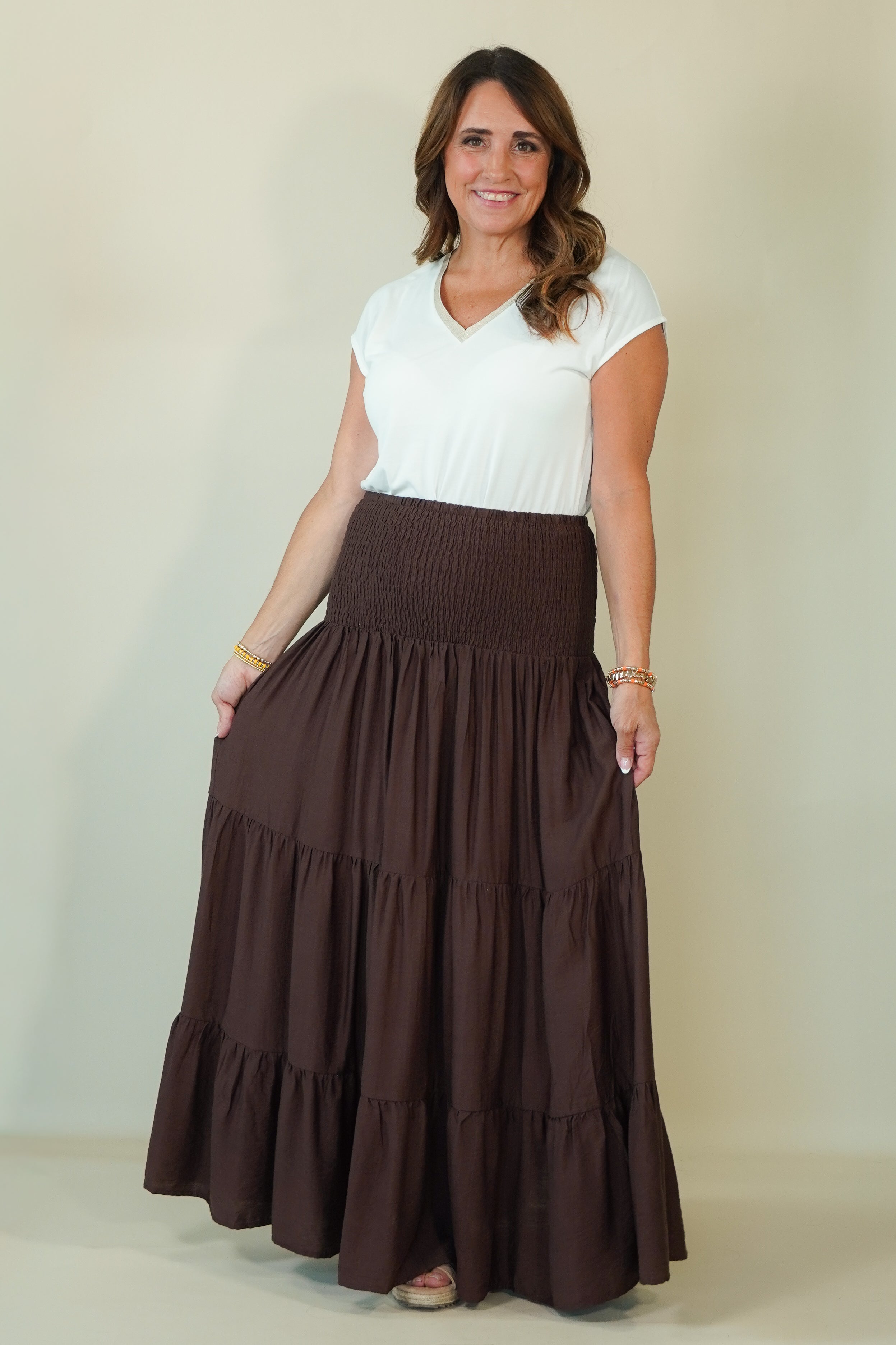 A woman modeling a white short-sleeve V-neck top and a high-waisted, dark brown tiered maxi skirt with side pockets, standing against a plain beige studio background.