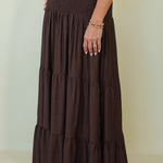 A woman modeling a white short-sleeve collared top paired with a long, dark brown tiered skirt against a plain beige studio background.