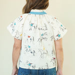 A woman viewed from the back wearing a cream-colored short-sleeve blouse with a blue collar and an all-over equestrian print of horses and riders, paired with blue jeans, against a plain beige background.