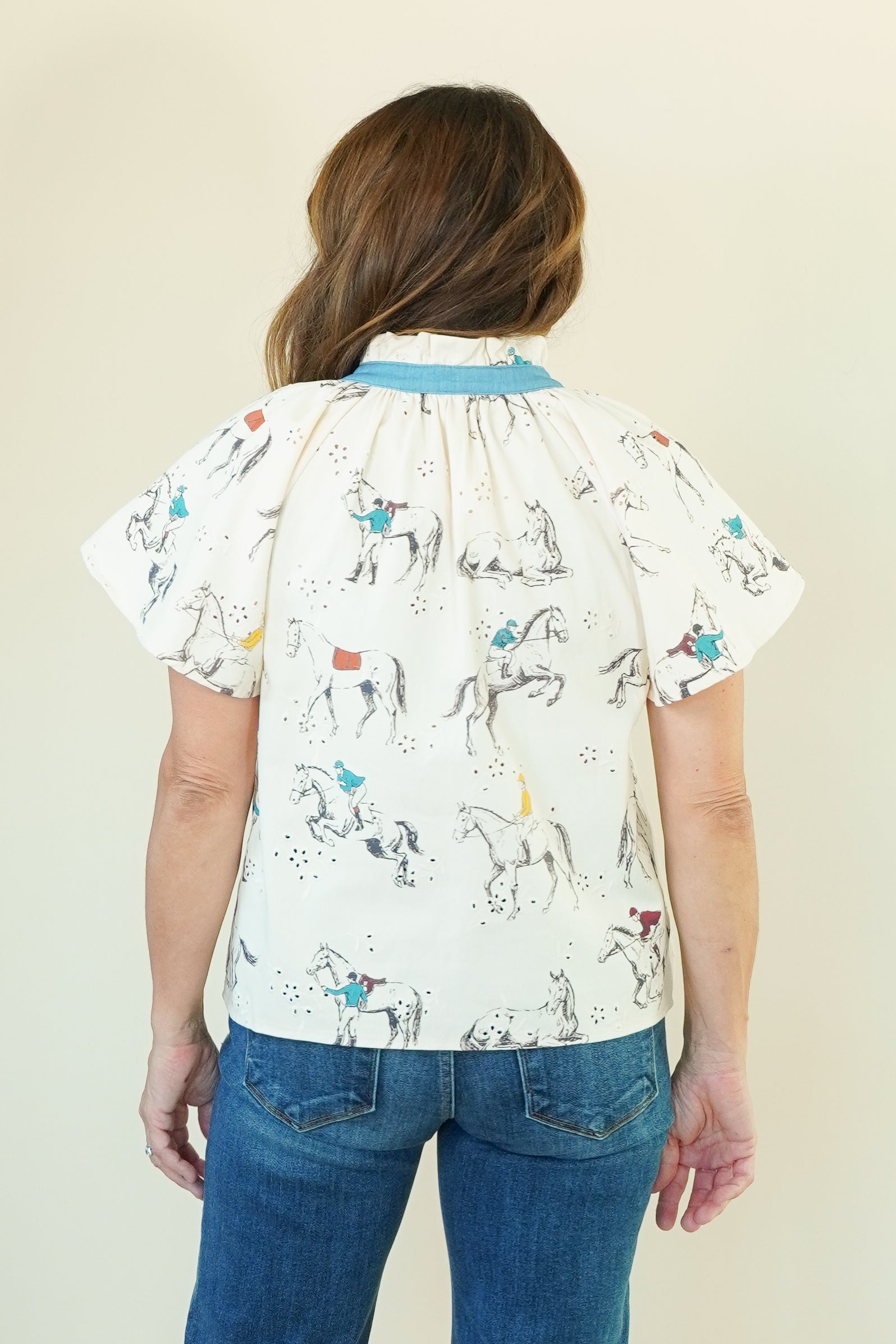 A woman viewed from the back wearing a cream-colored short-sleeve blouse with a blue collar and an all-over equestrian print of horses and riders, paired with blue jeans, against a plain beige background.