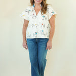 A woman modeling a white short-sleeve button-up blouse with a colorful cartoon animal print, paired with blue jeans, standing against a plain cream background.