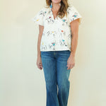 A woman modeling a white short-sleeve button-up shirt with a colorful cowboy and horse print, paired with blue jeans, standing against a plain cream-colored background.
