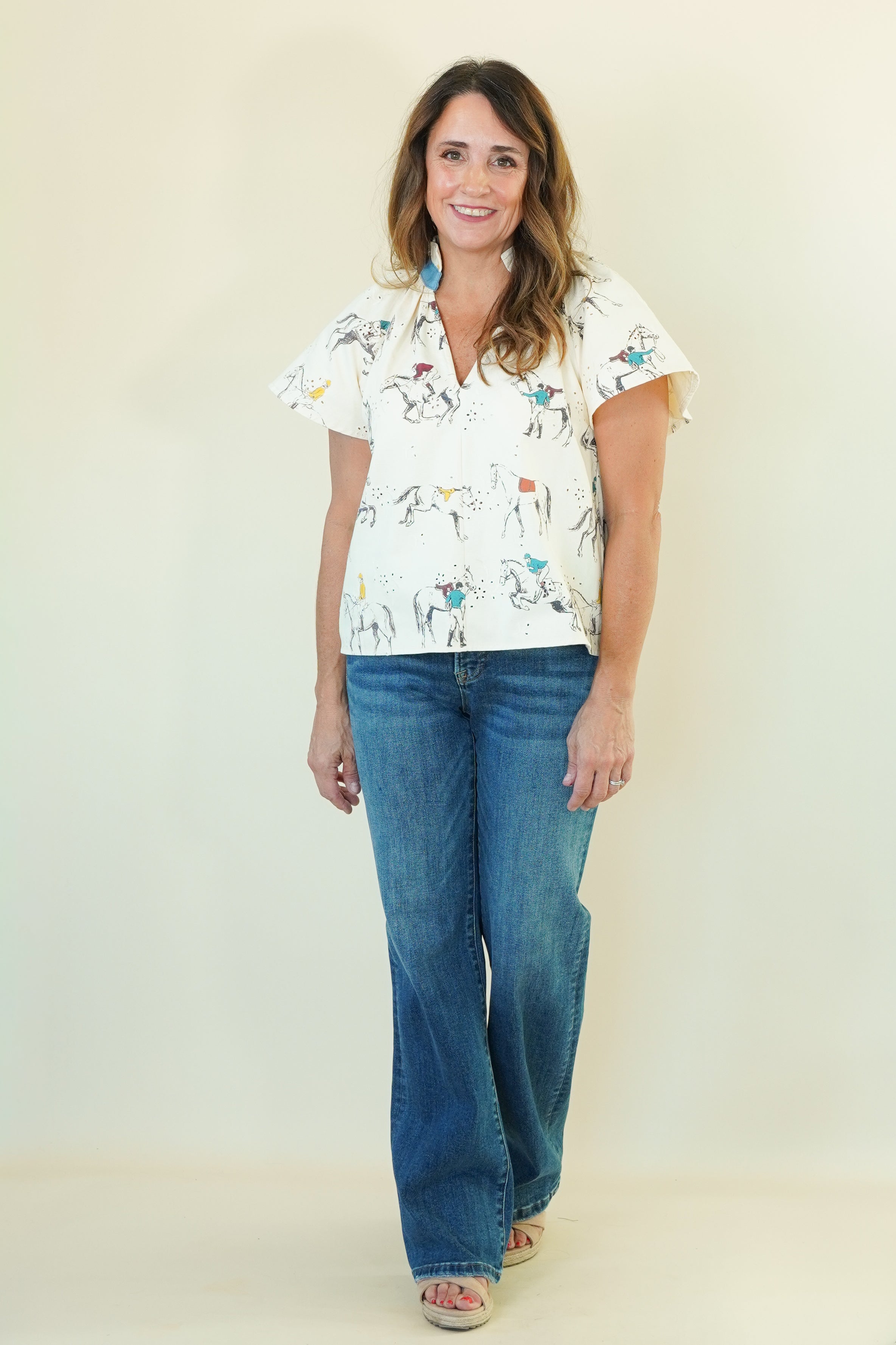 A woman modeling a white short-sleeve button-up shirt with a colorful cowboy and horse print, paired with blue jeans, standing against a plain cream-colored background.