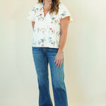 A woman modeling a white short-sleeve button-up shirt with a colorful cowboy and cattle print, paired with blue jeans and beige slide sandals, standing against a plain cream background.