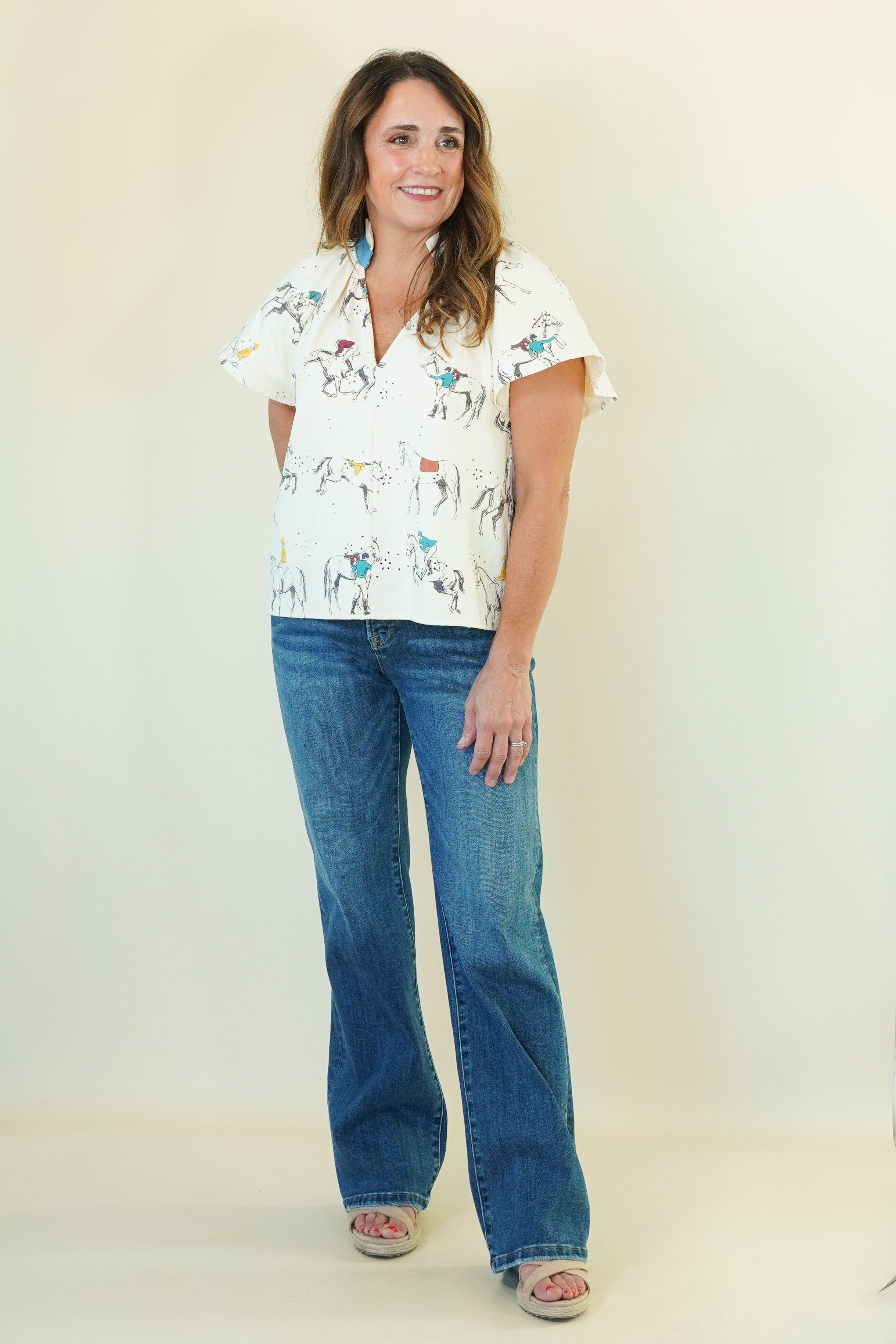 A woman modeling a white short-sleeve button-up shirt with a colorful cowboy and cattle print, paired with blue jeans and beige slide sandals, standing against a plain cream background.