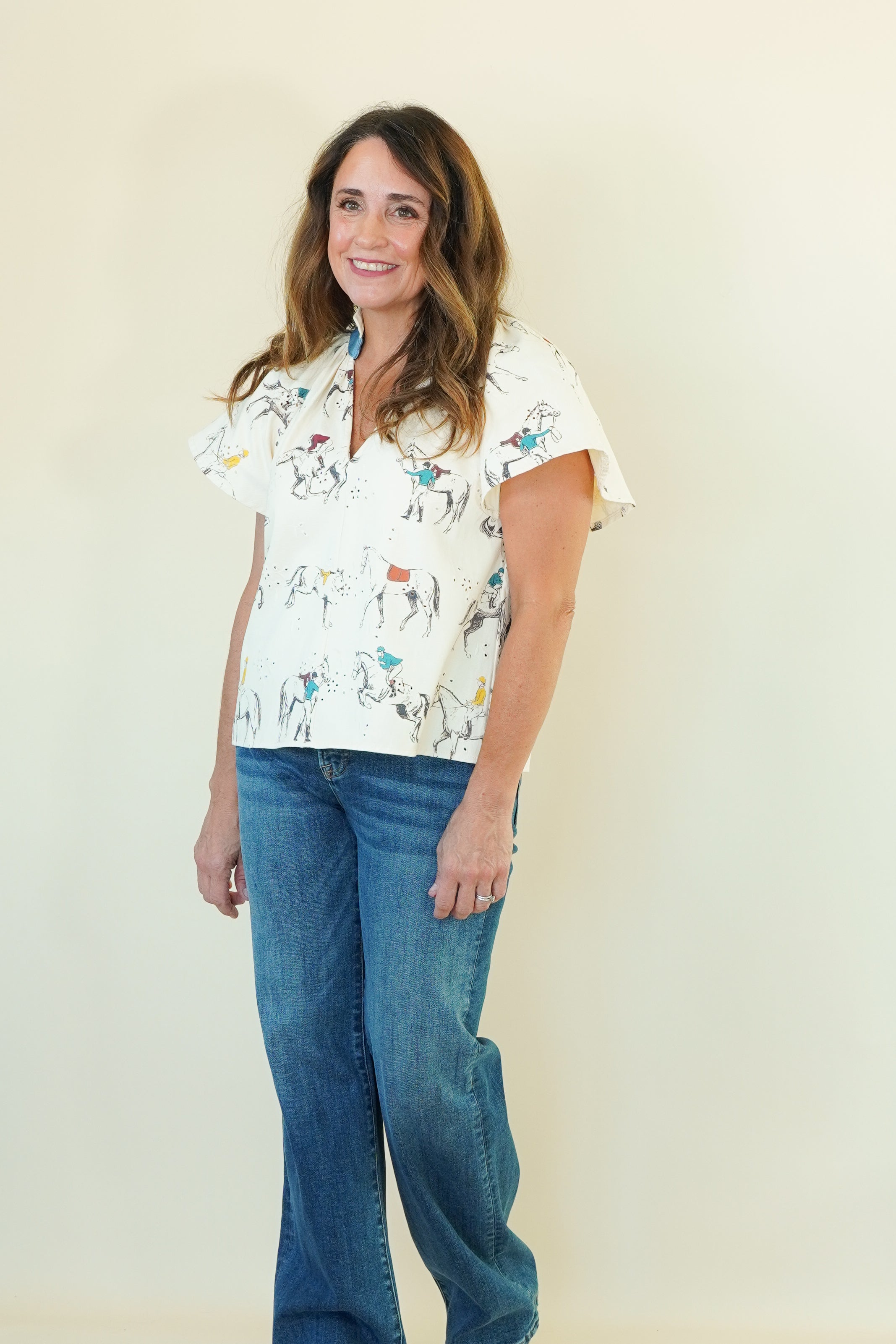 A woman modeling a short-sleeve button-up shirt with a white base and a colorful cartoon animal print, paired with blue jeans, standing against a plain cream background.