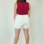 A woman shown from behind wearing a sleeveless burgundy top and white shorts, standing against a plain light beige studio background.