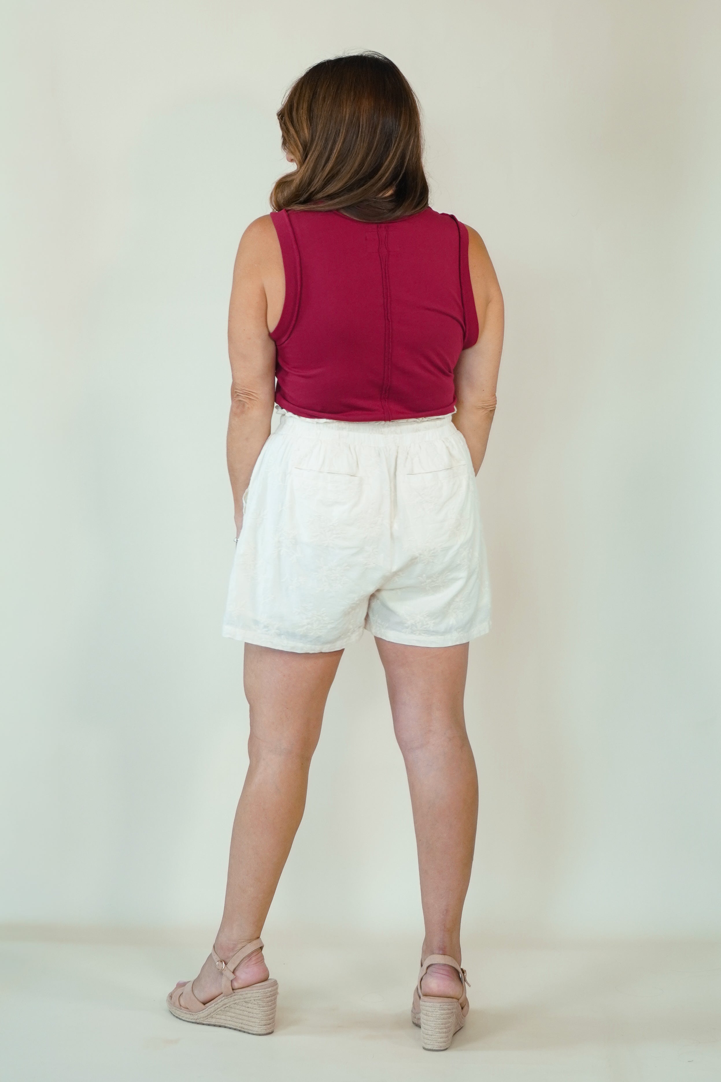 A woman shown from behind wearing a sleeveless burgundy top and white shorts, standing against a plain light beige studio background.