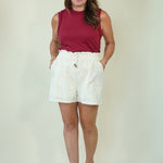 A woman modeling a sleeveless burgundy top and white shorts against a plain light beige studio background.