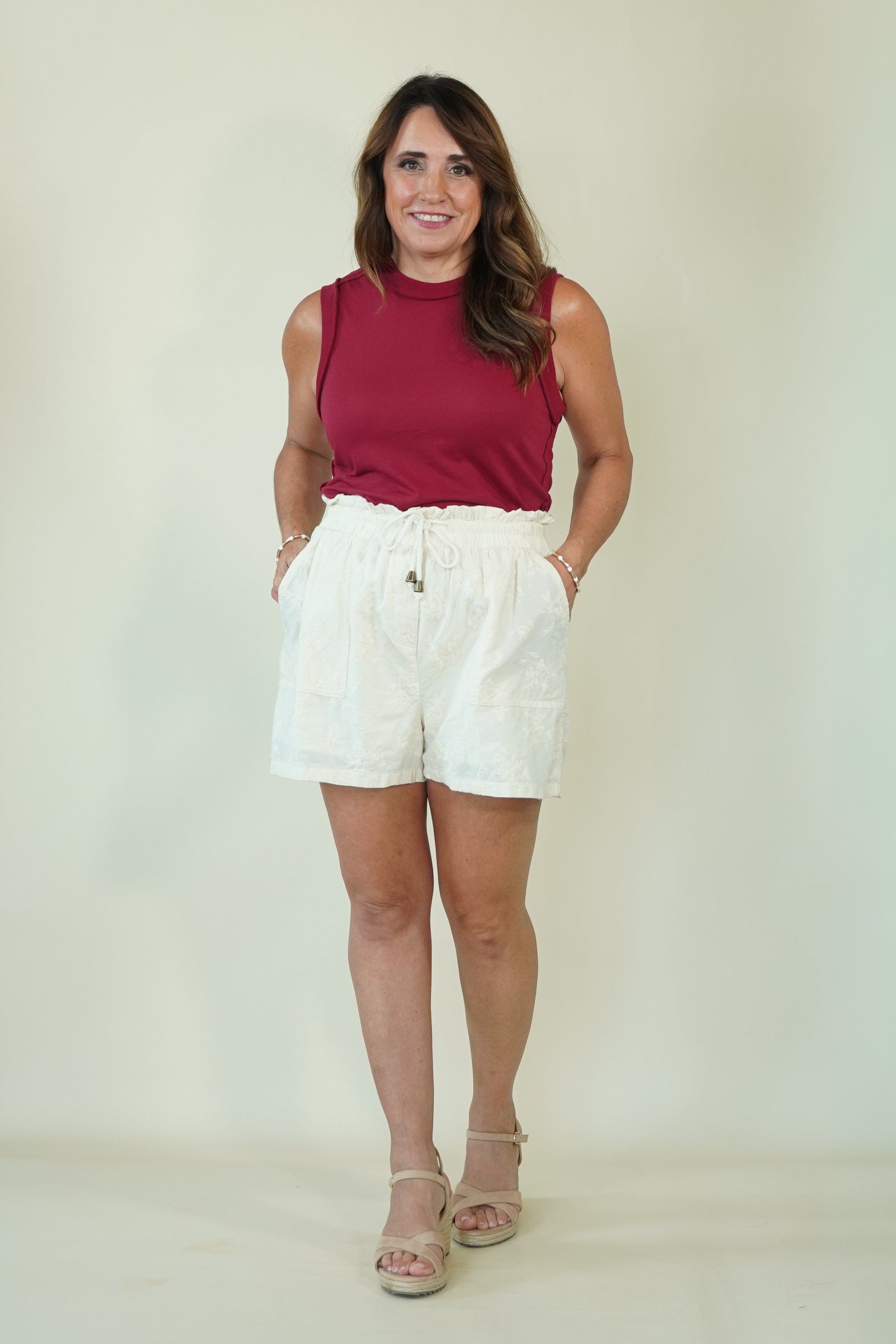 A woman modeling a sleeveless burgundy top and white shorts against a plain light beige studio background.