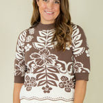 A woman wearing a brown and white knit sweater with a floral and leaf pattern, paired with white pants, standing against a plain beige background.