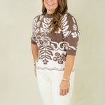 A woman modeling a brown and white floral patterned knit sweater with three-quarter sleeves, paired with white pants, against a plain beige studio background.