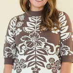 A woman modeling a brown and white floral patterned mock-neck knit sweater with elbow-length sleeves, paired with off-white pants, against a plain cream studio background.