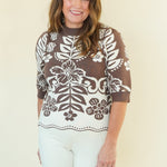 A woman modeling a brown and white patterned knit top with three-quarter sleeves, paired with white pants, standing against a plain cream-colored studio background.