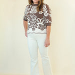 A woman modeling a brown and white floral patterned short-sleeve top with white pants, standing against a plain cream-colored studio background.
