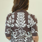 A woman viewed from behind wearing a brown short-sleeve knit top with a large white floral and leaf pattern, paired with white flared pants, against a plain off-white studio background.