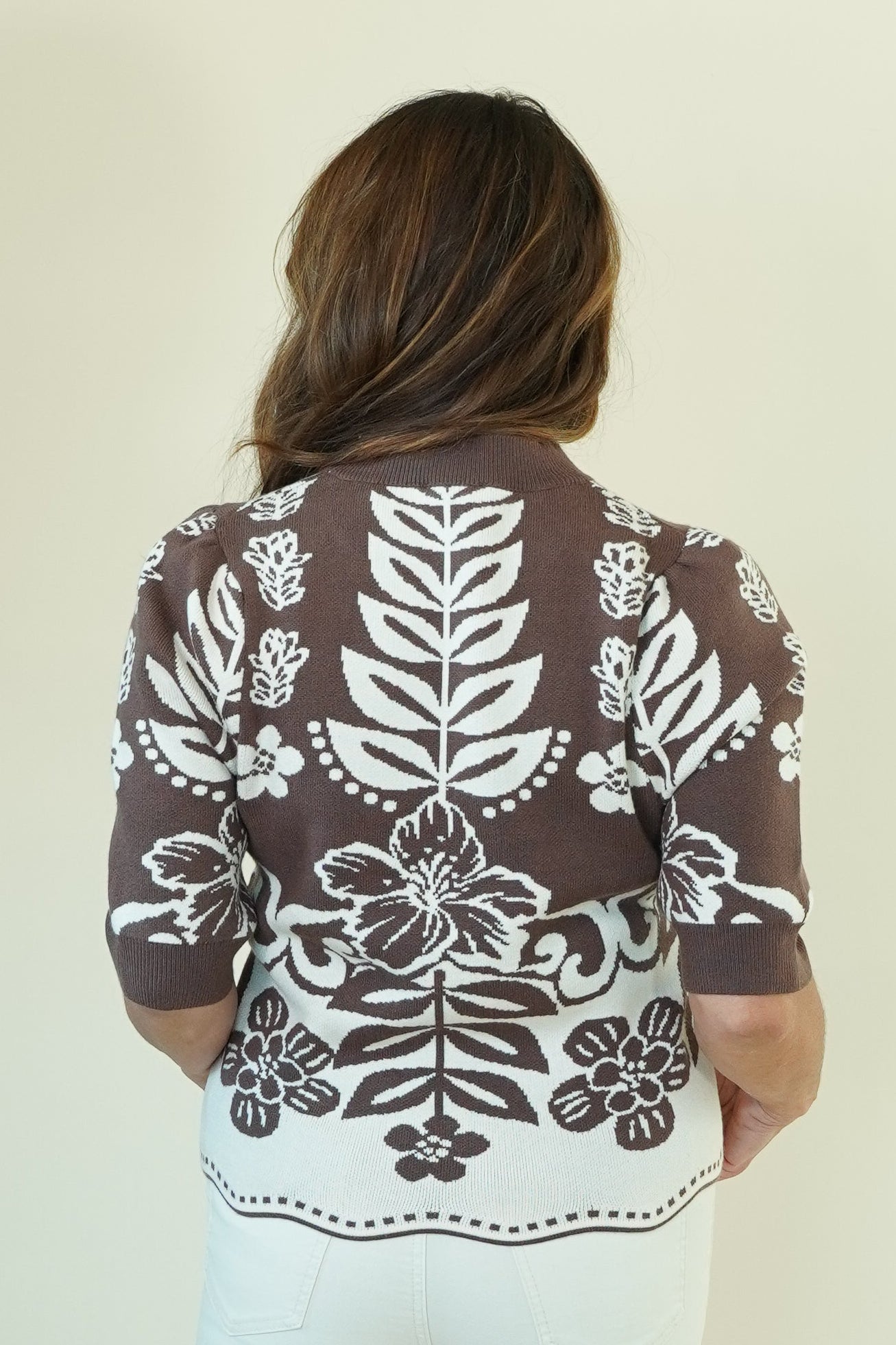 A woman viewed from behind wearing a brown short-sleeve knit top with a large white floral and leaf pattern, paired with white flared pants, against a plain off-white studio background.