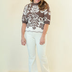 A woman modeling a brown and white floral patterned short-sleeve knit top with white pants, standing against a plain cream-colored studio background.